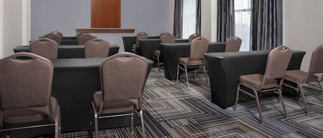 A conference room with multiple tables and chairs, a wooden accent wall, and large windows with curtains.
