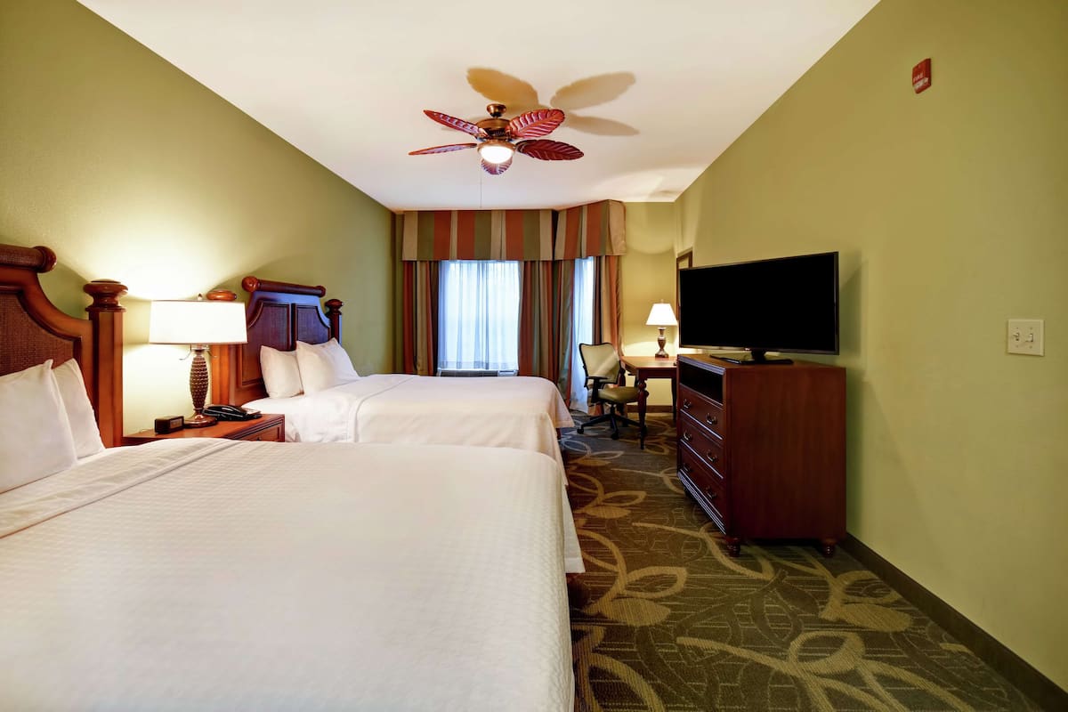 A hotel room with two beds, a ceiling fan, a television, a desk with a chair, and a dresser.
