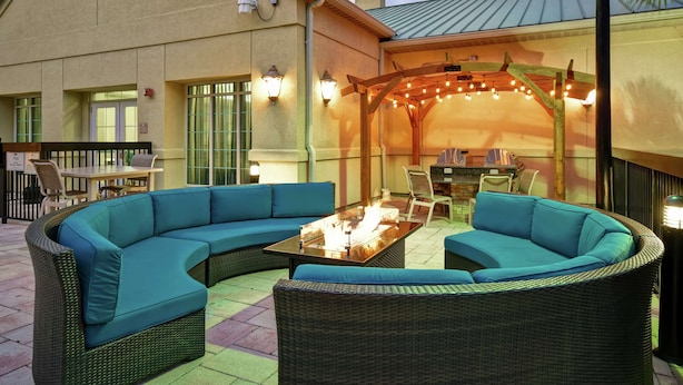 Eine Terrasss mit blauer, gebogener Couch, einem Glastisch mit Feuerstelle und einer Holz-Pergola mit Lichterketten.