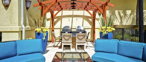 Une terrasse extérieure dotée d’une pergola, de deux canapés bleus, d’une table à plateau en verre et d’un barbecue.