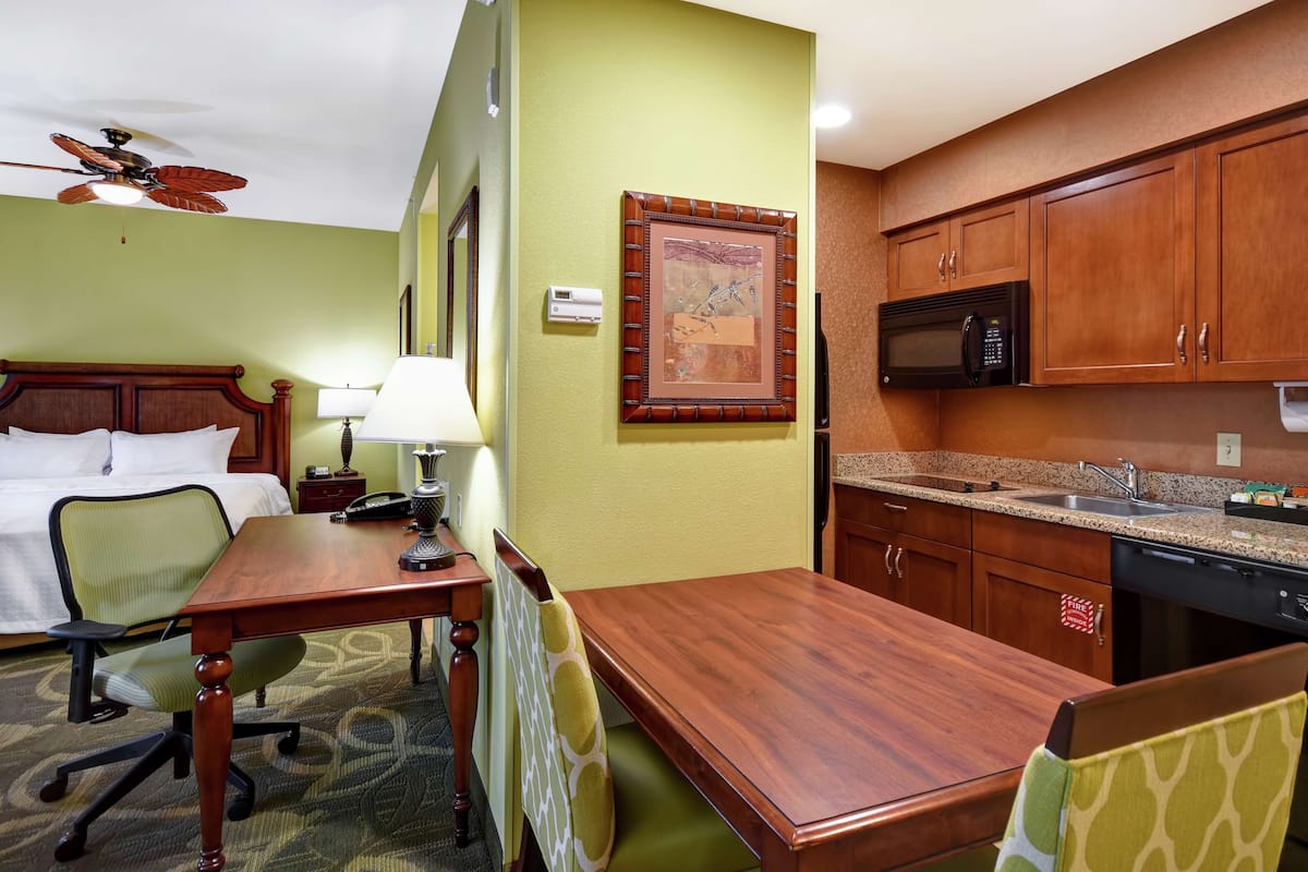 A hotel room with a wooden dining table, a bed with a green headboard, a ceiling fan, a microwave, a sink, and a framed picture on the wall.