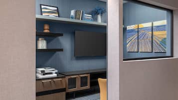 A modern office space with a flat-screen TV, shelves with books and decorative items, a printer, and a window with a view of fields.