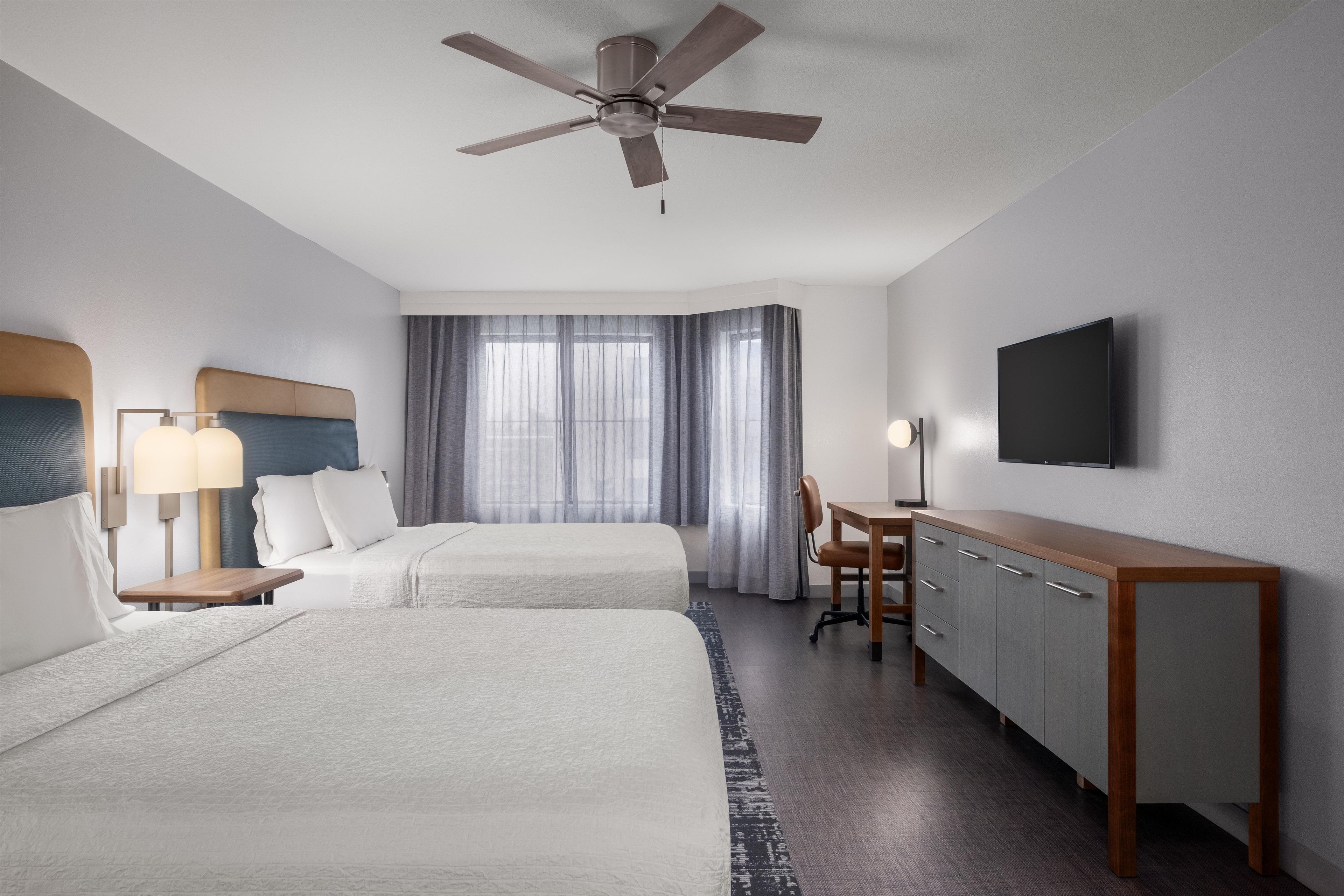 Habitación de hotel con dos camas, un escritorio, una silla, un televisor y un ventilador de techo.