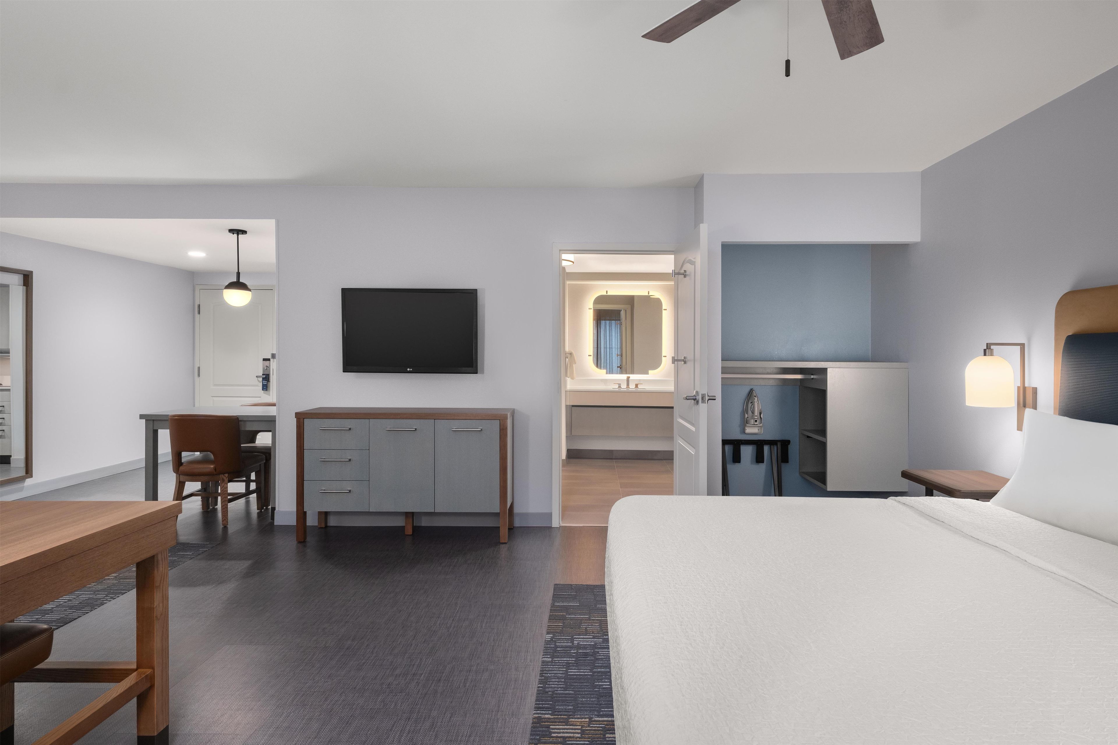 A hotel room with a bed, a desk, a chair, a TV, a kitchenette, and a ceiling fan.