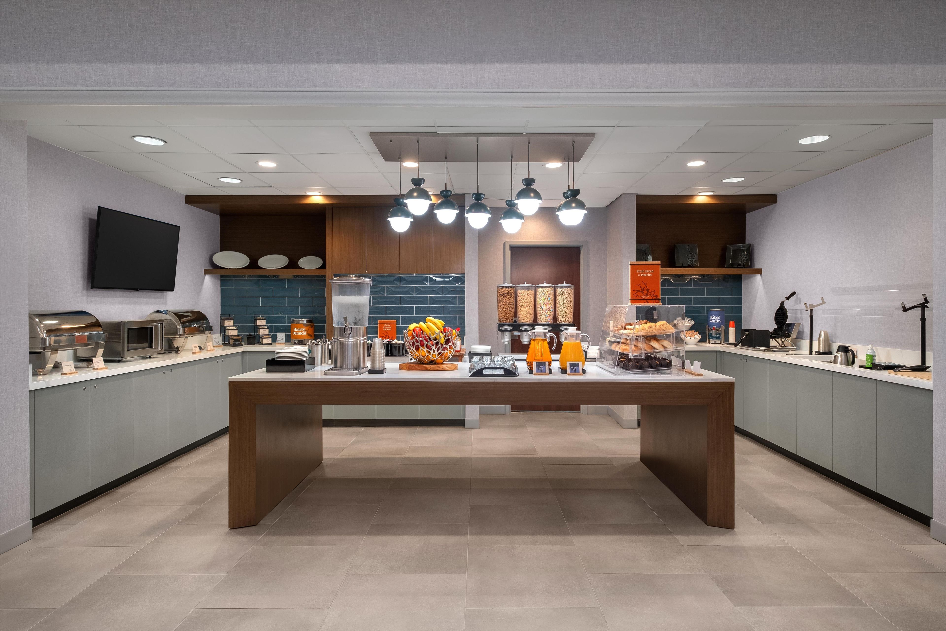 Uma área de café da manhã moderna com uma mesa central, balcão de café e diversos distribuidores de alimentos e bebidas.