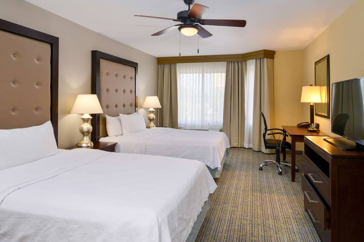 Ein Hotelzimmer mit zwei Betten, einem Schreibtisch, einem Sessel, einem Fernseher und einem Deckenventilator.