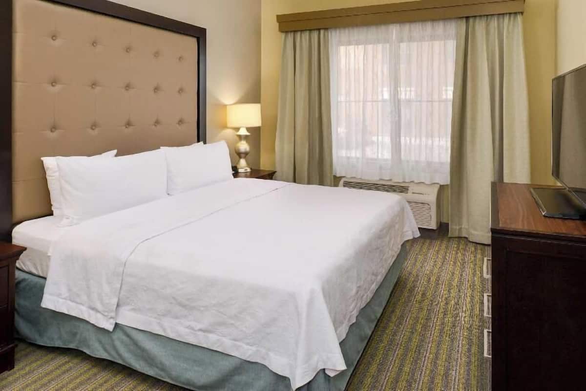 Ein Hotelzimmer mit einem großen Bett, einem Fernseher, einer Lampe und einem Fenster mit Vorhängen.