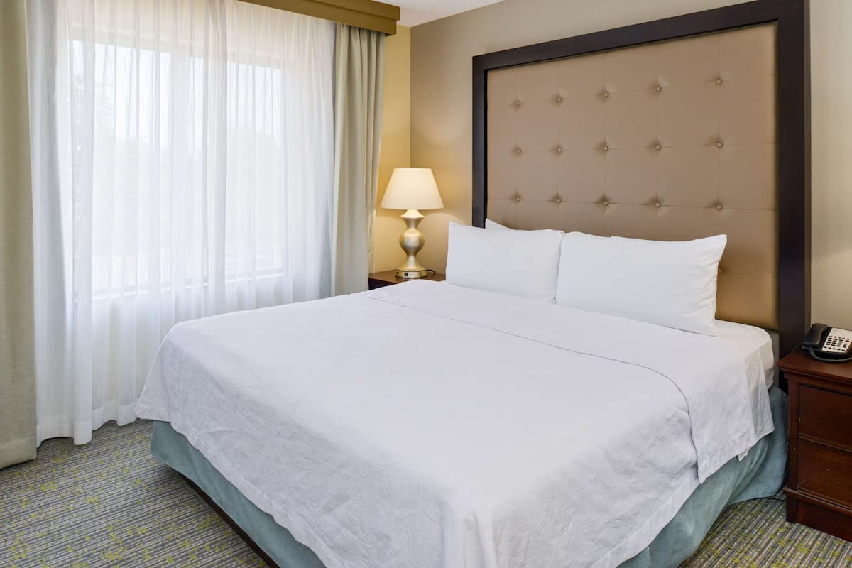 Ein Hotelzimmer mit einem großen Bett, einer Nachttischlampe, einem Telefon und einem Fenster mit Vorhängen.