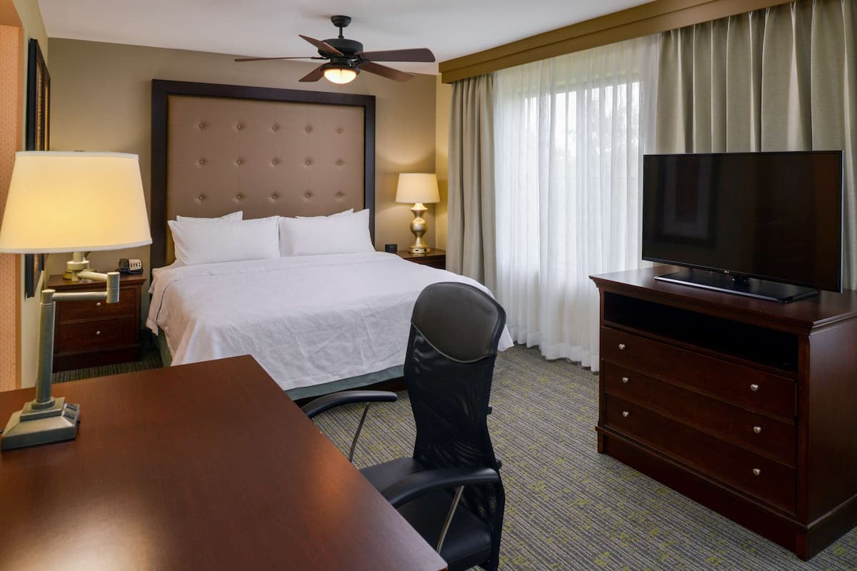 Ein Hotelzimmer mit Bett, Schreibtisch, Stuhl, Fernseher, Deckenventilator und Vorhängen.