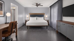 A hotel room with a bed, desk, chair, bedside tables, lamp, TV, and a ceiling fan.