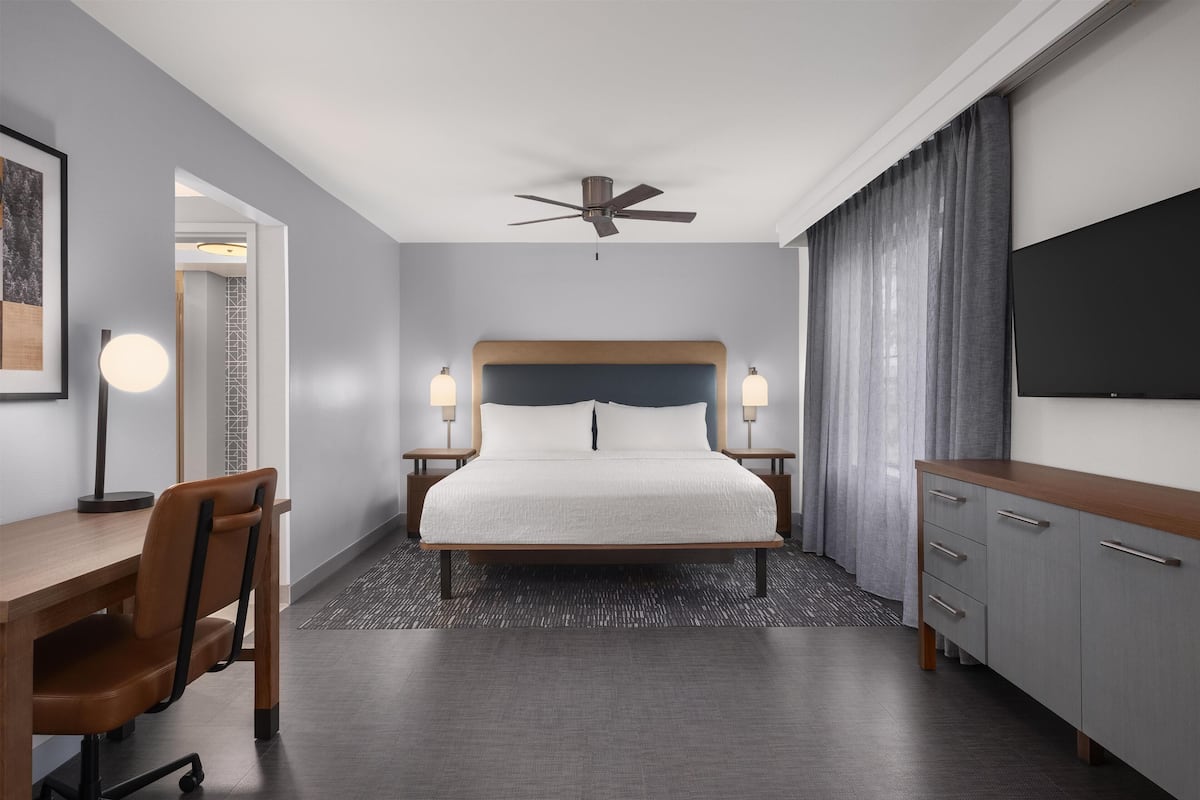 Ein Hotelzimmer mit Bett, Schreibtisch, Stuhl, Nachttischen, Lampe, Fernseher und Deckenventilator.
