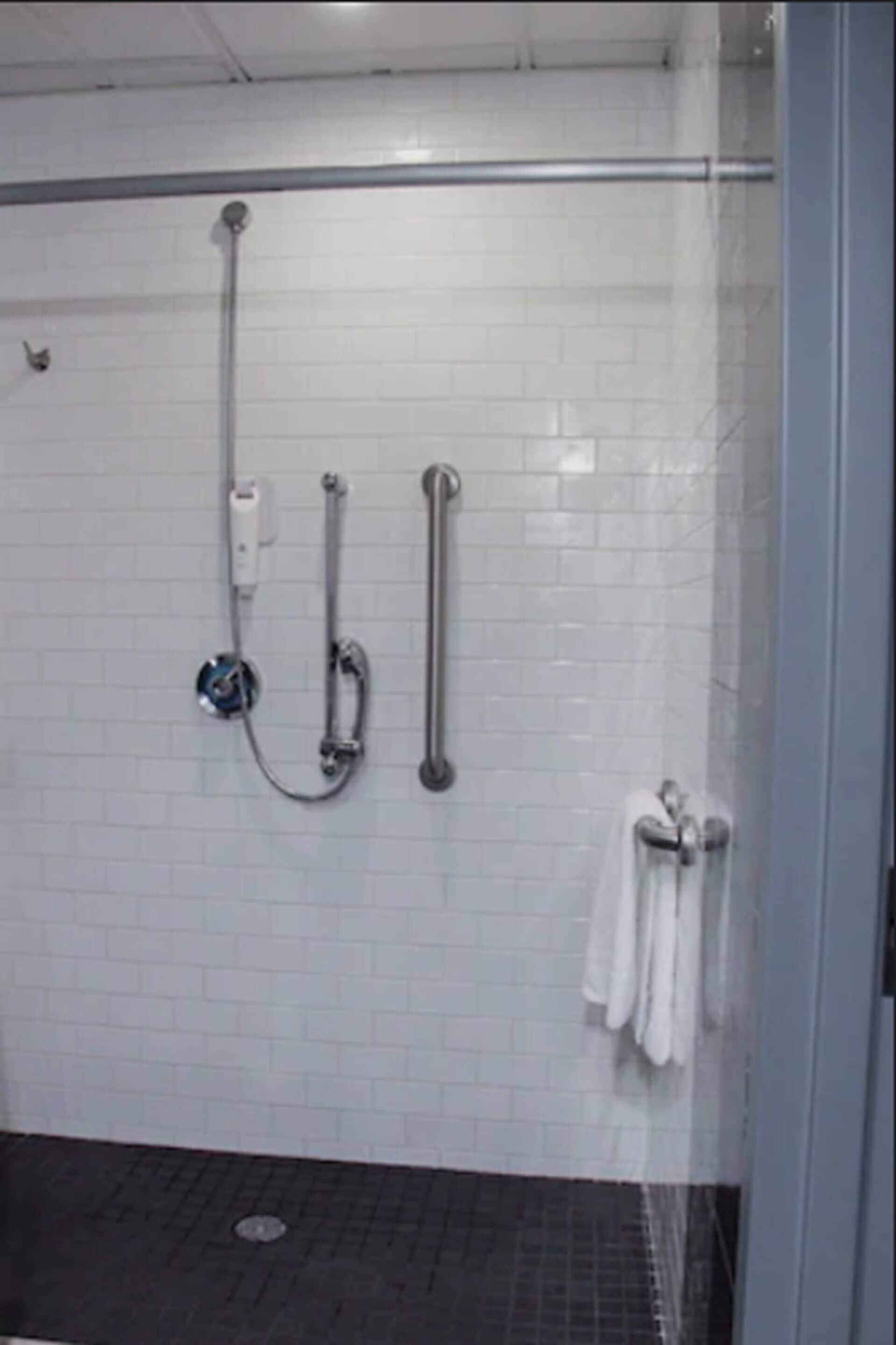 Un espace douche avec des murs recouverts de carreaux blancs, une porte en verre et une barre d’appui en métal.