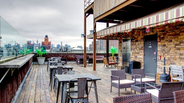 A rooftop bar with outdoor seating, a city skyline view, and a brick building with a striped awning. - The Ridge Hotel (New York)