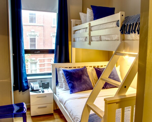 A bunk bed with white bedding and blue pillows, a bedside table with a phone and a lamp, and a window with curtains.