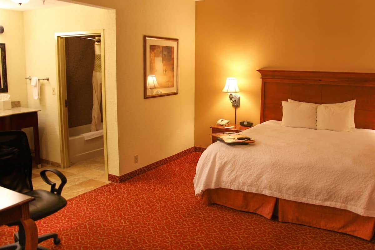 A hotel room with a bed, a desk, a chair, a nightstand, a lamp, and a bathroom visible through an open door.