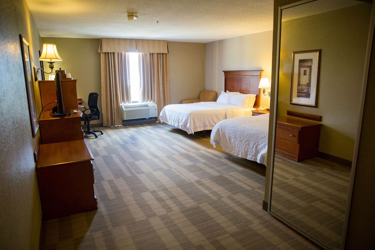 A hotel room with two beds, a desk with a chair, a dresser, a mirror, and a window with curtains.