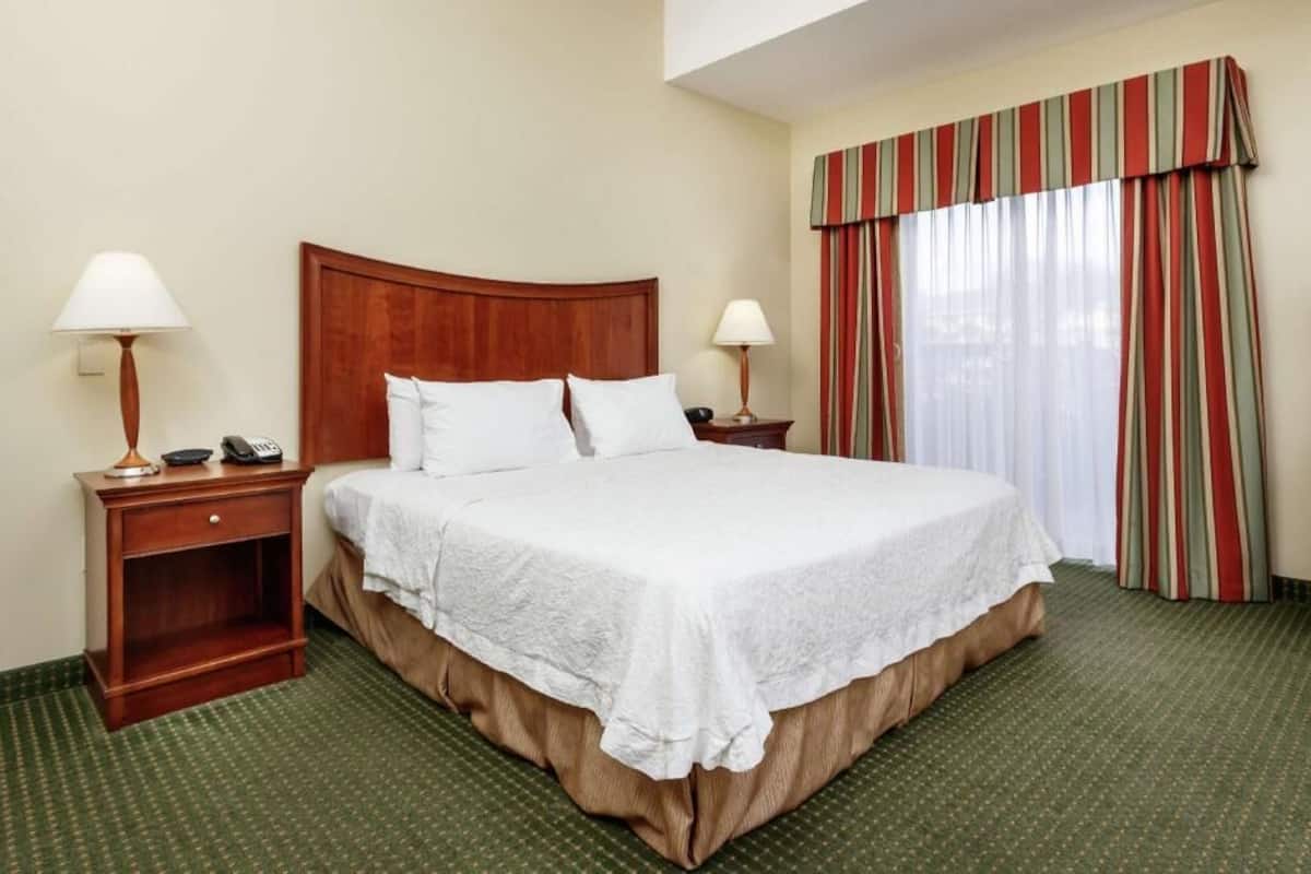 Habitación de hotel con una cama grande, dos mesitas de noche con lámparas, un cabecero de madera y una ventana con cortinas a rayas.
