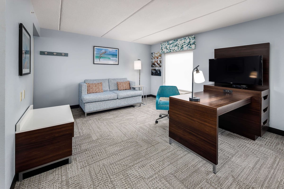 Habitación de hotel con sofá gris, escritorio con televisor, lámpara y ventana con vistas.