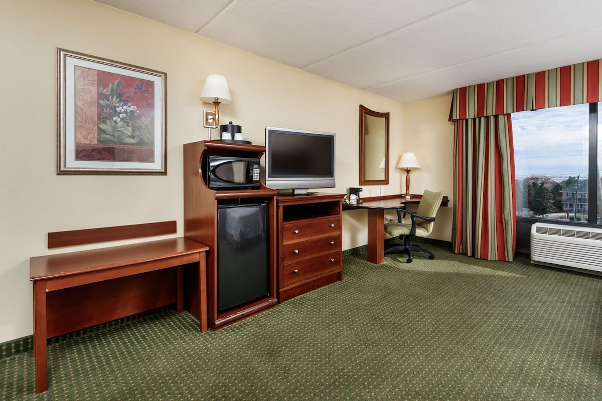 Habitación de hotel con escritorio, televisión, microondas, mini refrigerador, una silla, un ventanal con cortinas y un cuadro en la pared.