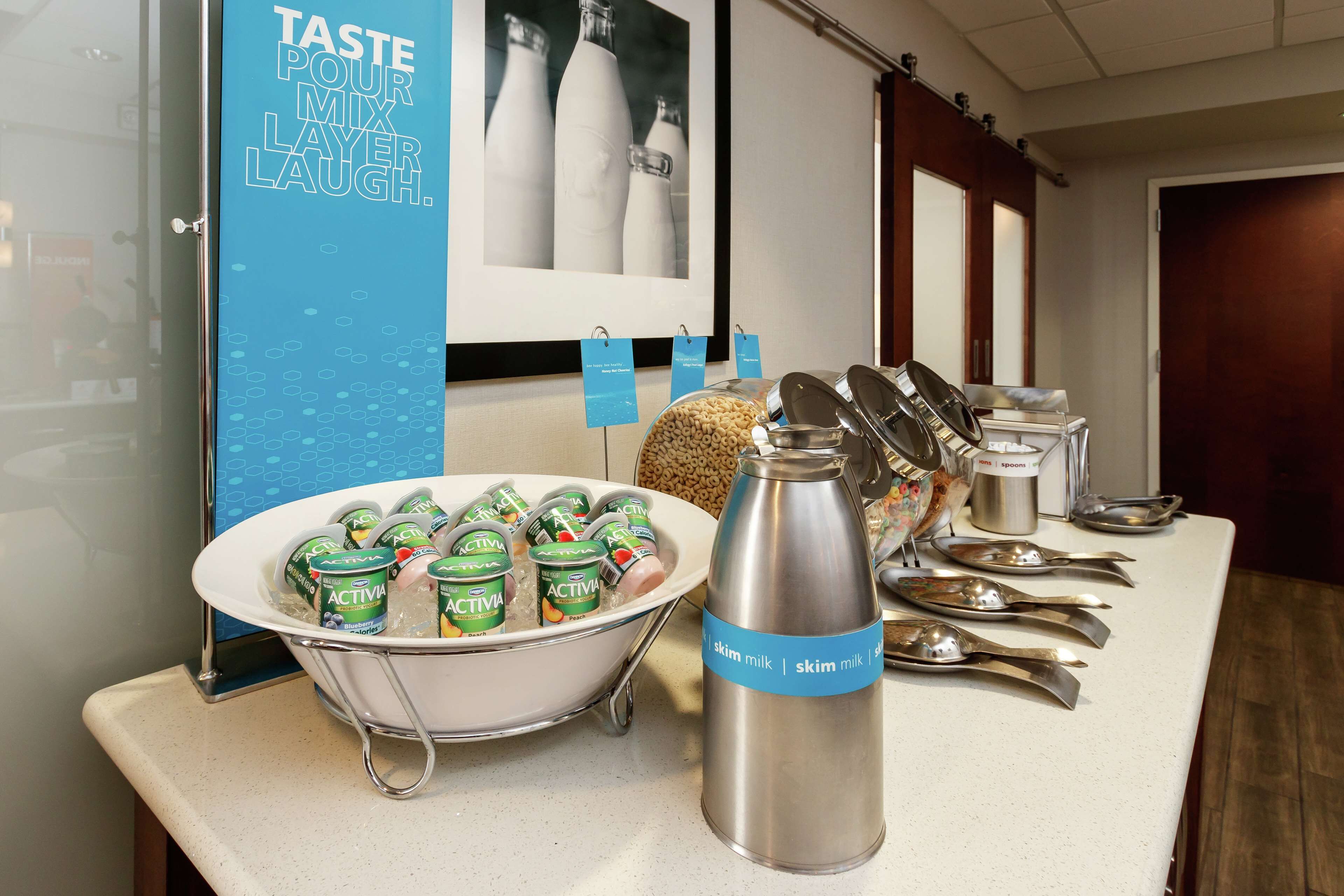 A buffet table with yogurt cups, a stainless steel milk bottle, and silver spoons.