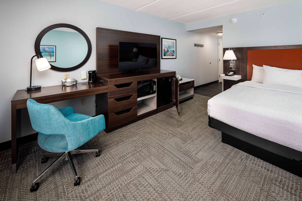Una habitación de hotel con cama, escritorio con televisor, silla y mesita de noche con lámpara.
