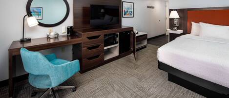Una habitación de hotel con cama, escritorio con televisor, silla y mesita de noche con lámpara.