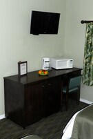 Un escritorio de madera oscura con un microondas blanco, una cafetera y un bol de fruta. Un televisor de pantalla plana está montado en la pared sobre el escritorio.