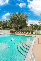 A clear blue pool with a diving board, surrounded by lounge chairs and a white fence.