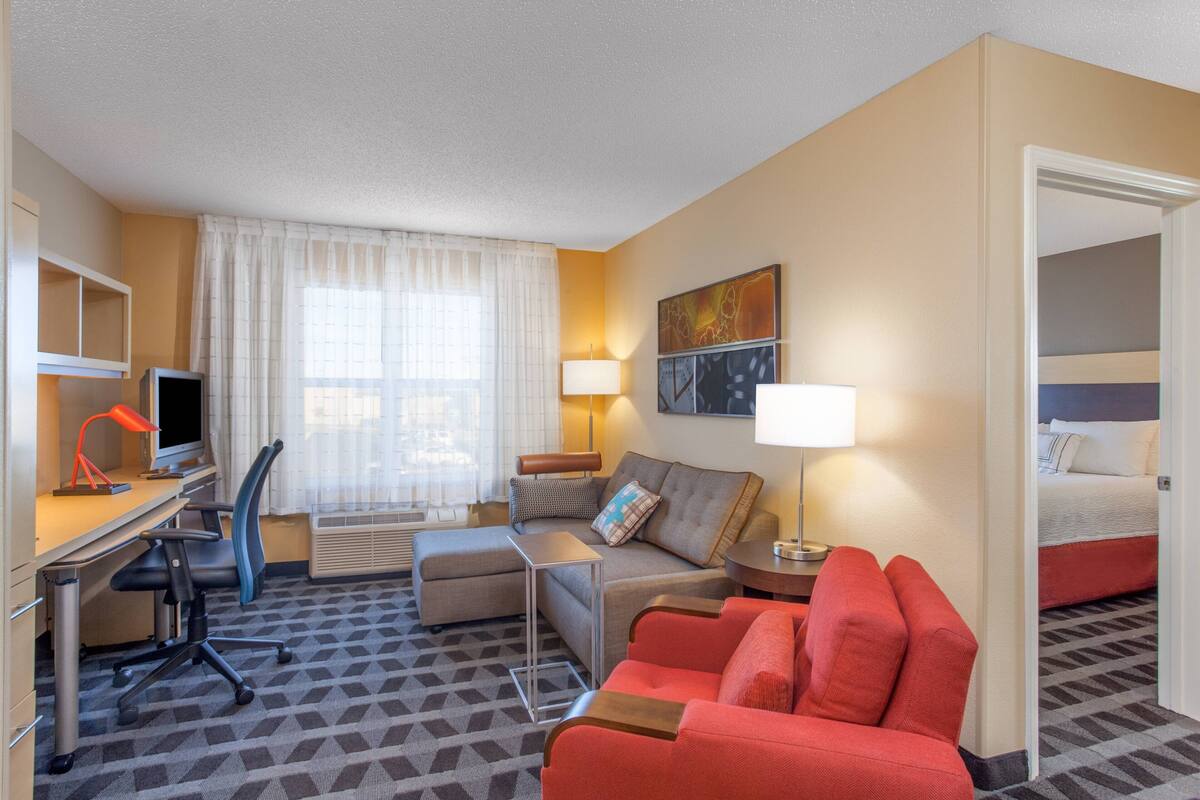 A hotel room with a desk, a sofa, a red armchair, a bed, and a painting on the wall.