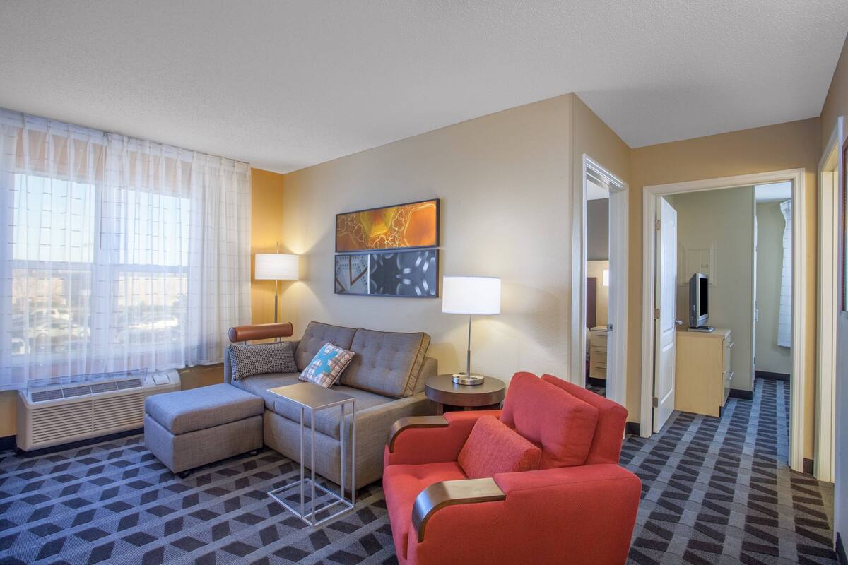 A hotel room with a sofa, ottoman, two lamps, a flat-screen TV, and a painting on the wall.