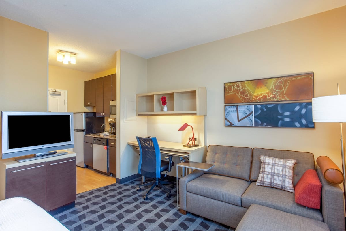 A hotel room with a TV, sofa, desk, chair, kitchenette, and a map on the wall.
