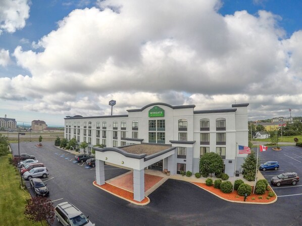 A modern hotel with a prominent 'WINGATE' sign, a parking lot with several cars, and a flagpole with flags.