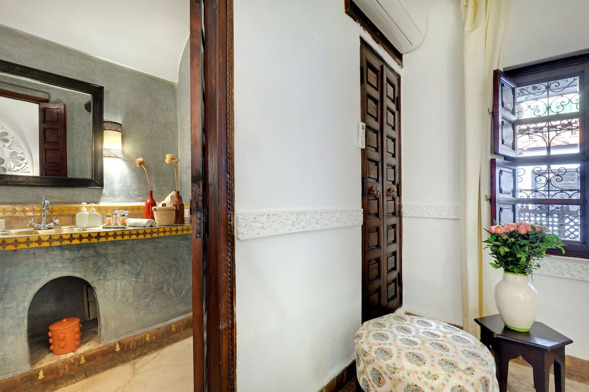 Une salle de bain avec un mur recouvert de carreaux, un miroir, un lavabo avec des articles de toilette, une petite fenêtre ornée de barreaux en fer décoratifs et un vase rempli de fleurs posé sur une table d’appoint.