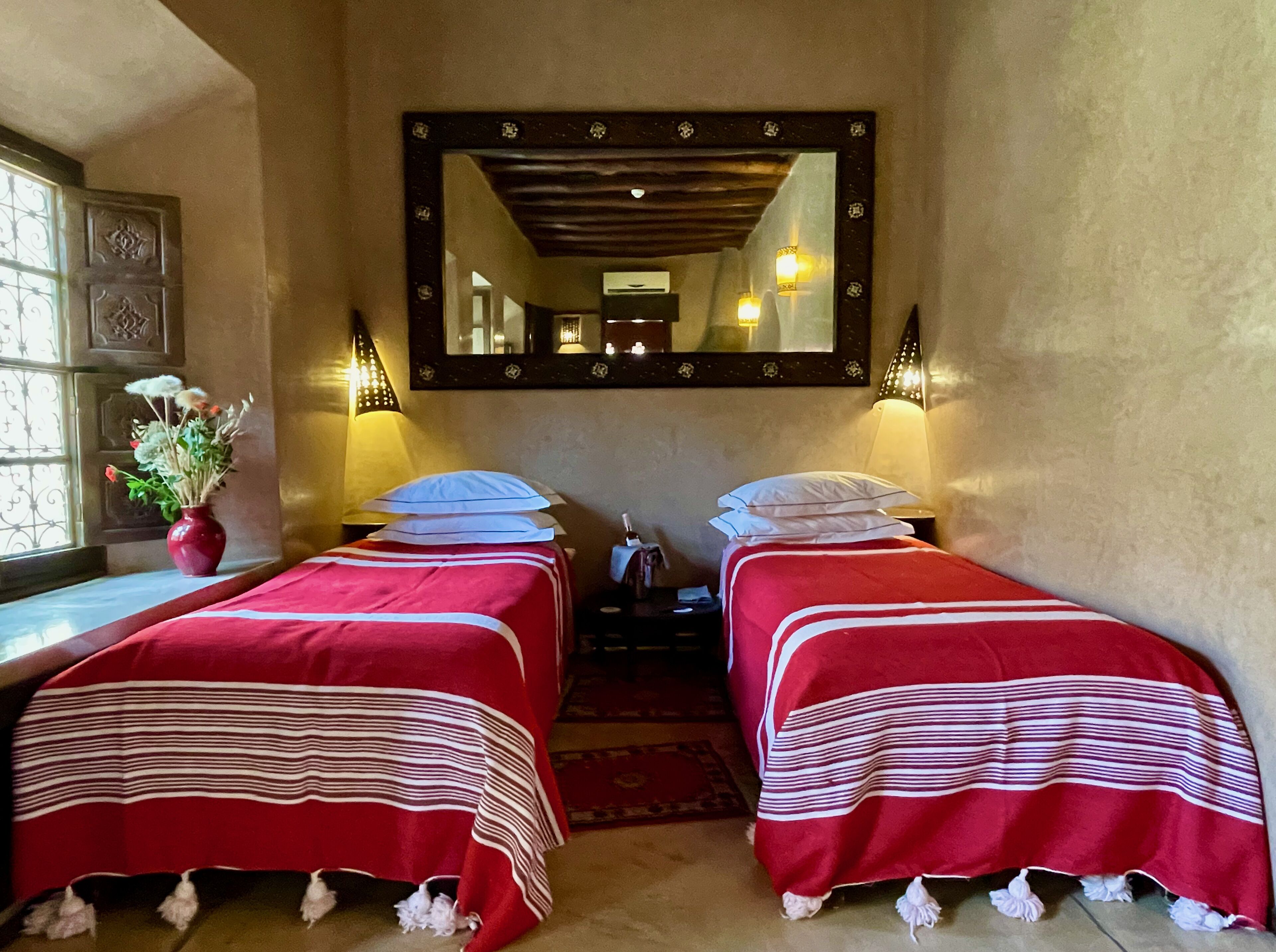 A hotel room with two single beds, a large mirror, bedside lamps, a vase with flowers, and a window with decorative iron bars.