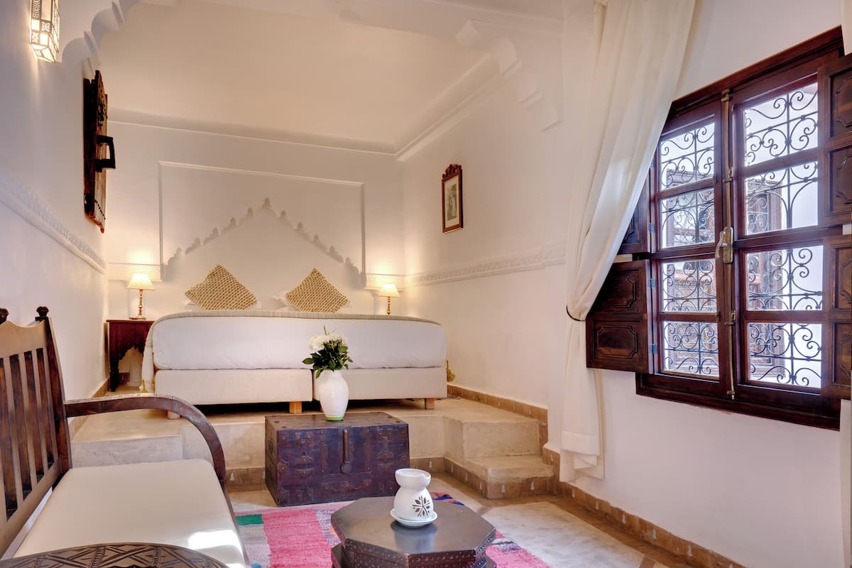 Une chambre à coucher avec un lit, des lampes de chevet, un vase avec des fleurs, une chaise en bois, une petite table et une fenêtre ornée de barreaux en fer décoratifs.