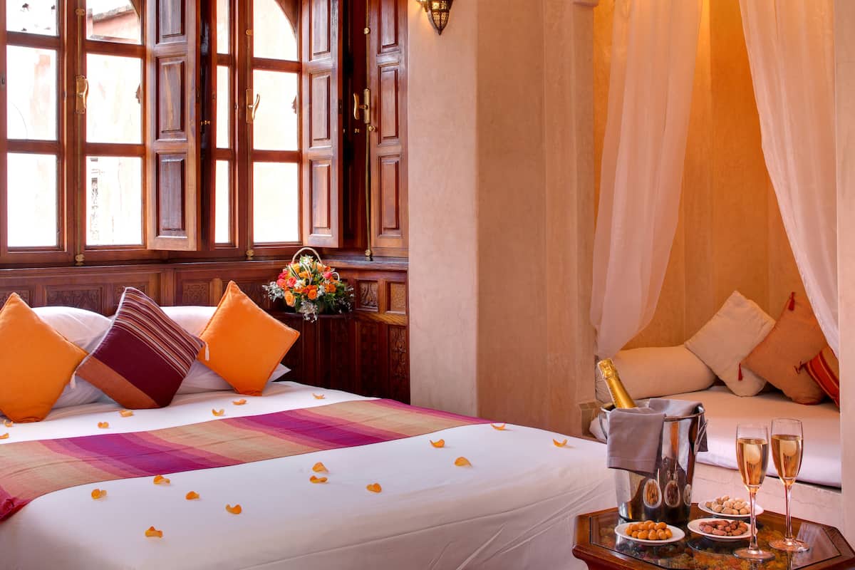 Une chambre d’hôtel avec un grand lit, une table de chevet sur laquelle se trouve une bouteille de champagne, deux verres à champagne et un plateau avec des noix et des chocolats.
