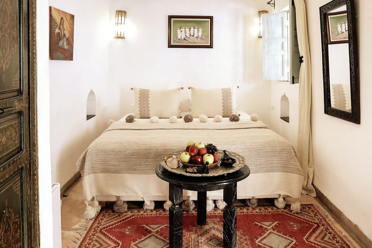 Une chambre à coucher avec un lit, une table de chevet sur laquelle se trouvent des fruits, un miroir et des lampes fixées au mur.