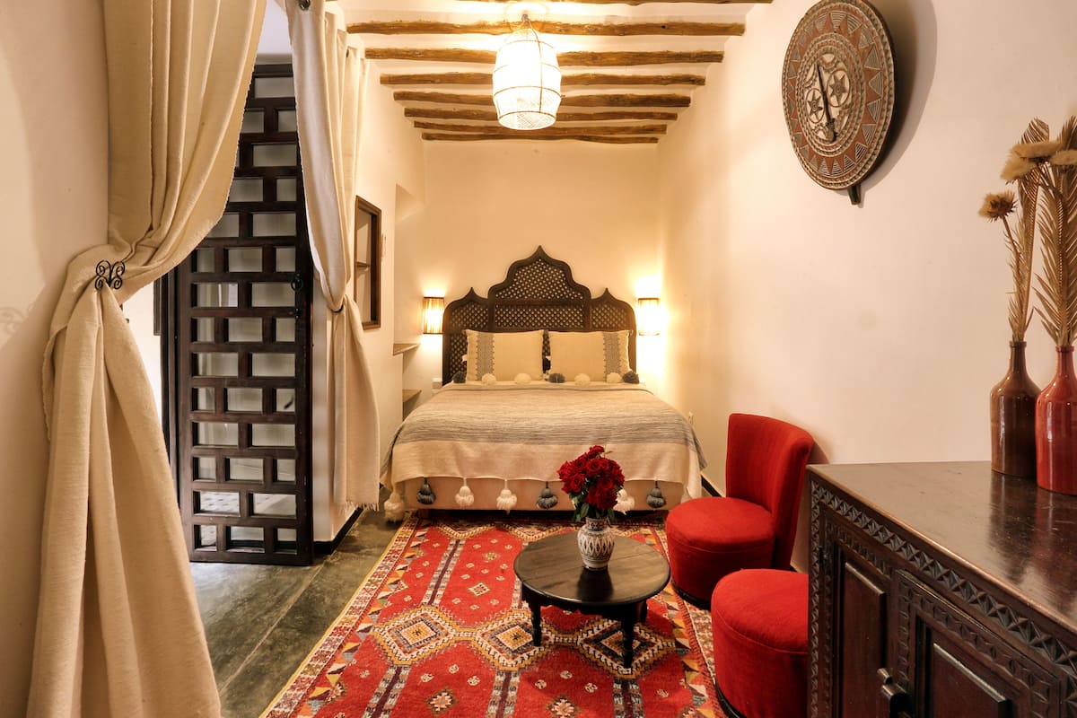 Une chambre à coucher avec un lit, un fauteuil rouge, une petite table sur laquelle est posé un vase de fleurs, et un élément décoratif mural.