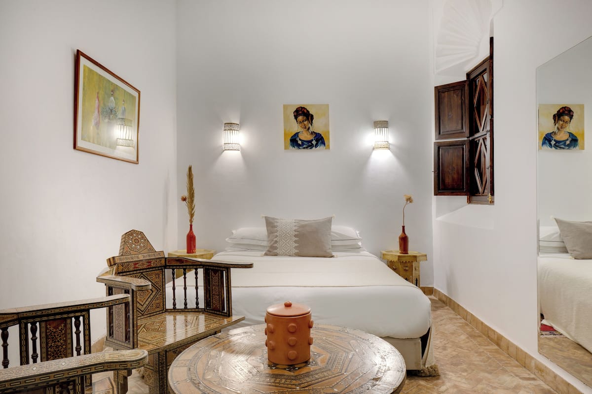 Une chambre d’hôtel comprenant un lit, deux lampes fixées au mur, un tableau, une chaise en bois, une petite table et un vase décoratif.