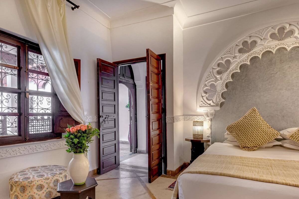 Une chambre à coucher avec un grand lit, une table de chevet avec une lampe, une fenêtre avec des rideaux et une porte donnant sur une autre pièce.