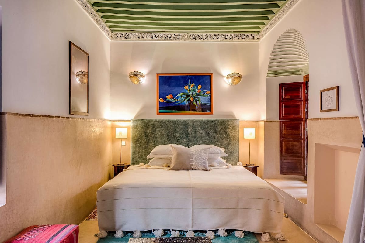 Une chambre d’hôtel avec un lit, un tableau, des appliques murales, un tapis à motifs et une lampe décorative.