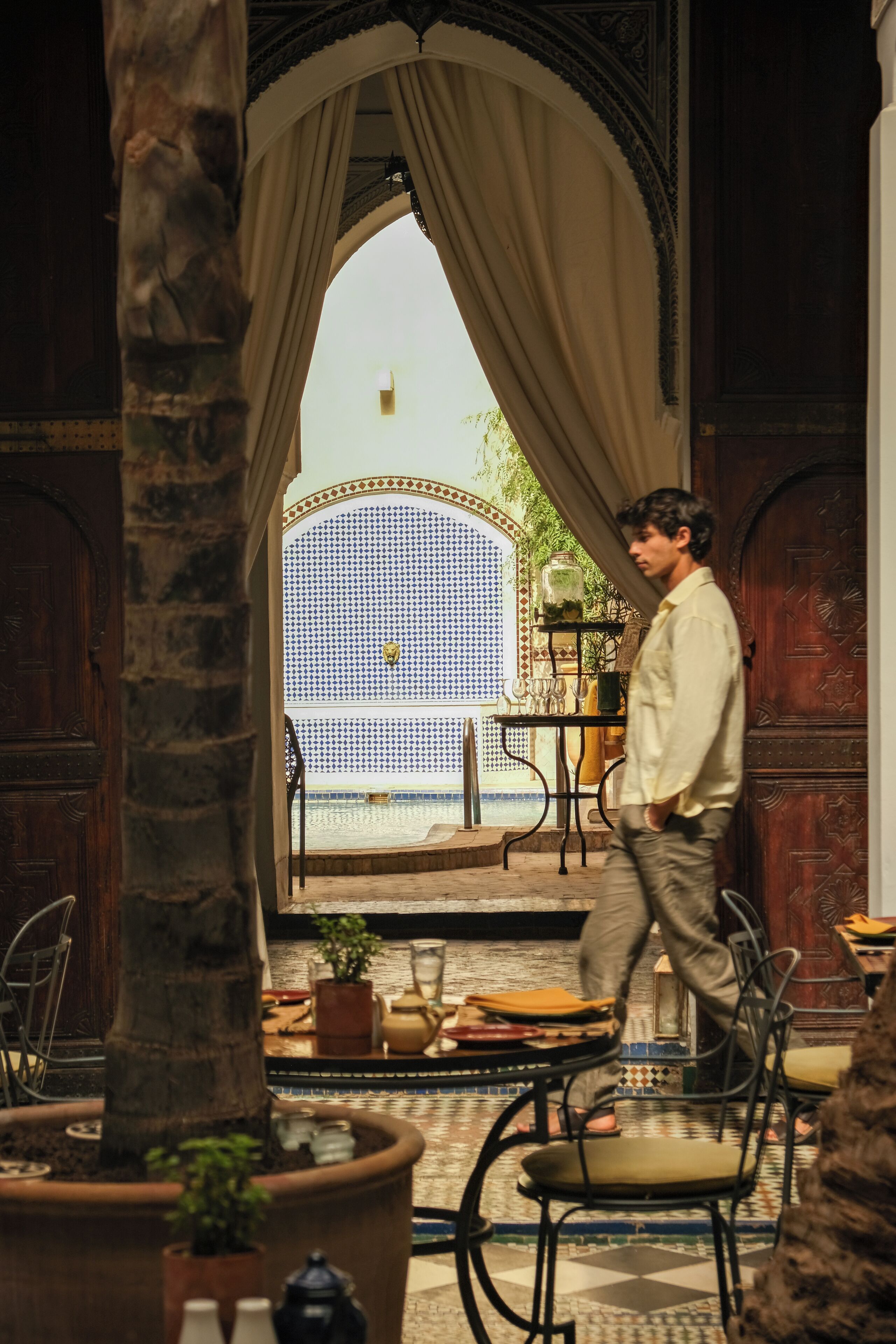 A man walking through an ornate doorway into a room with a tiled floor, a table with a potted plant, and a chair.