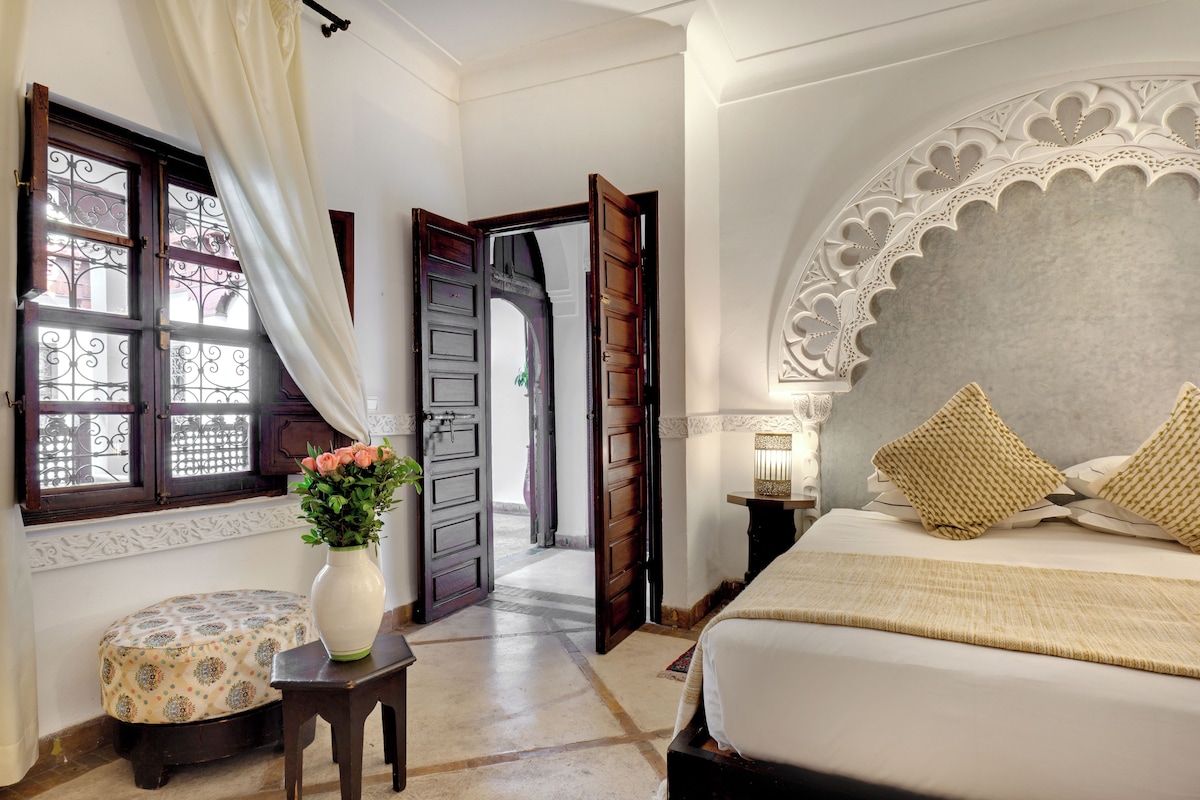 A bedroom with a bed, a nightstand, a lamp, a window with curtains, and a door leading to another room.