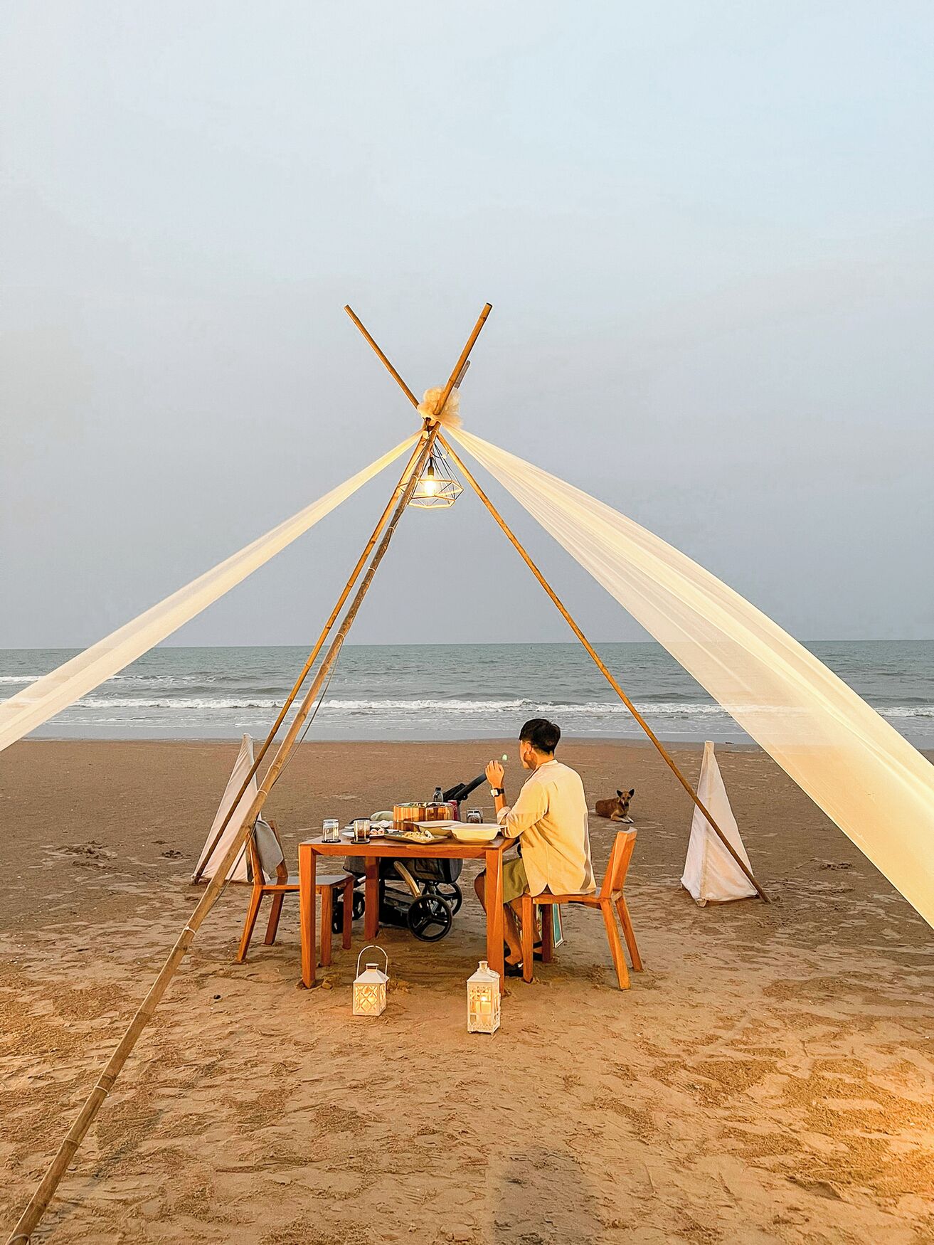 A beach scene with a tent set up on the sand, a table with food and drinks, and a dog nearby.
