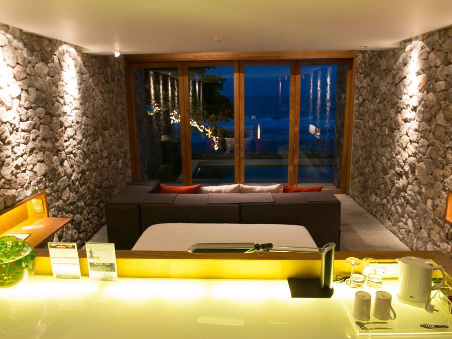 Habitación de hotel con pared de piedra, cama grande con almohadas, cabecero de madera, ventana de vidrio con vistas y mesita de noche con artículos de cortesía.