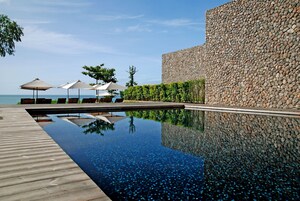 Un área de piscina moderna con terraza de madera, pared de piedra y sombrillas.