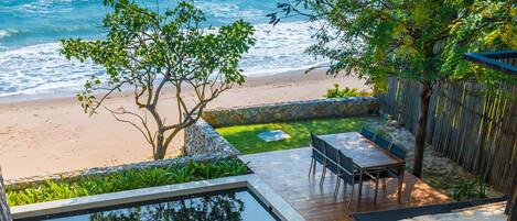 A wooden deck with a stone wall, a small pool, and a table with chairs overlooking a beach.