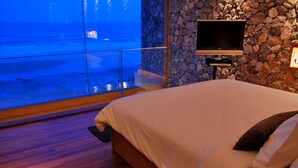 Un dormitorio con una cama grande, un televisor montado en una pared de piedra y vistas a la playa a través de una puerta de cristal.