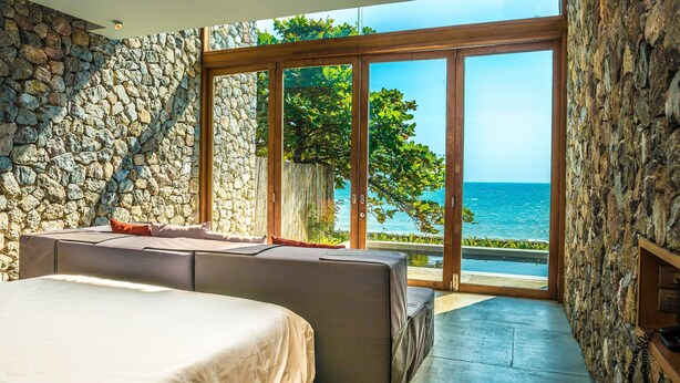 Un dormitorio con pared de piedra, cama, sofá y un amplio ventanal corredera que da a un balcón con vistas al mar.