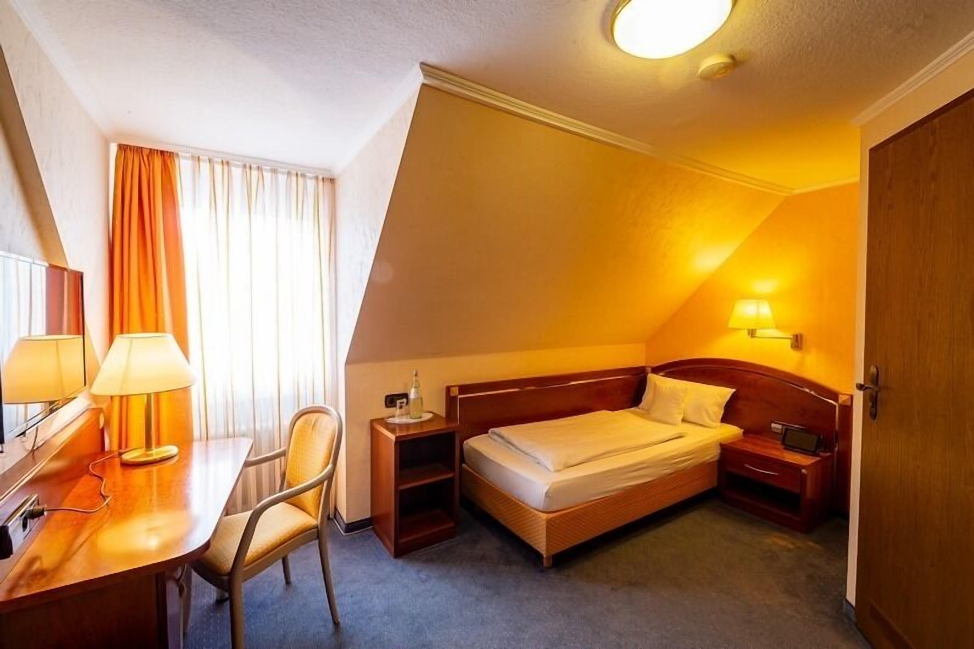 A hotel room with a bed, bedside table, lamp, chair, and a desk with a lamp.